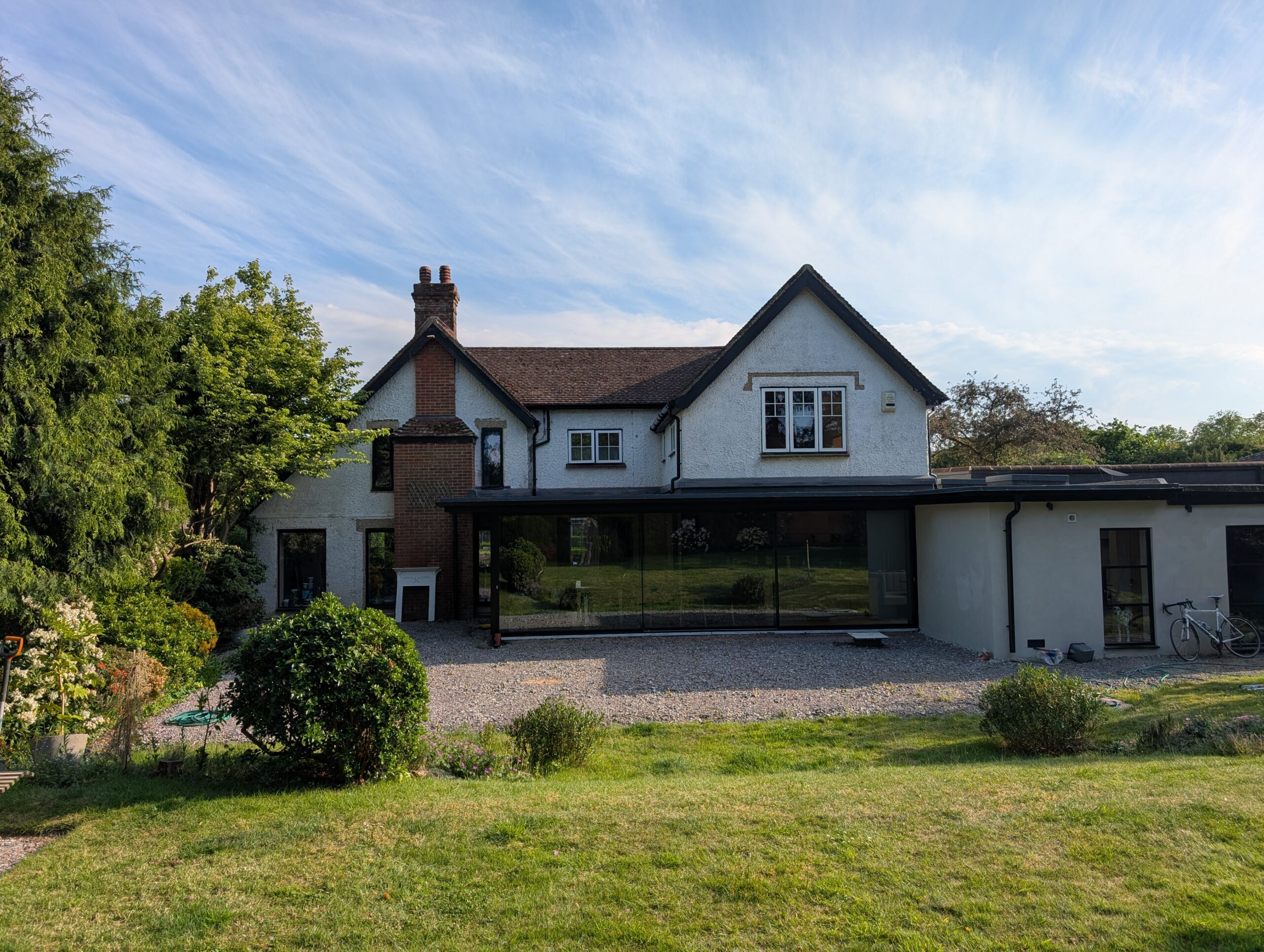 Two-story white house with a brick chimney, glass extension, and green garden under a blue sky with wispy clouds.