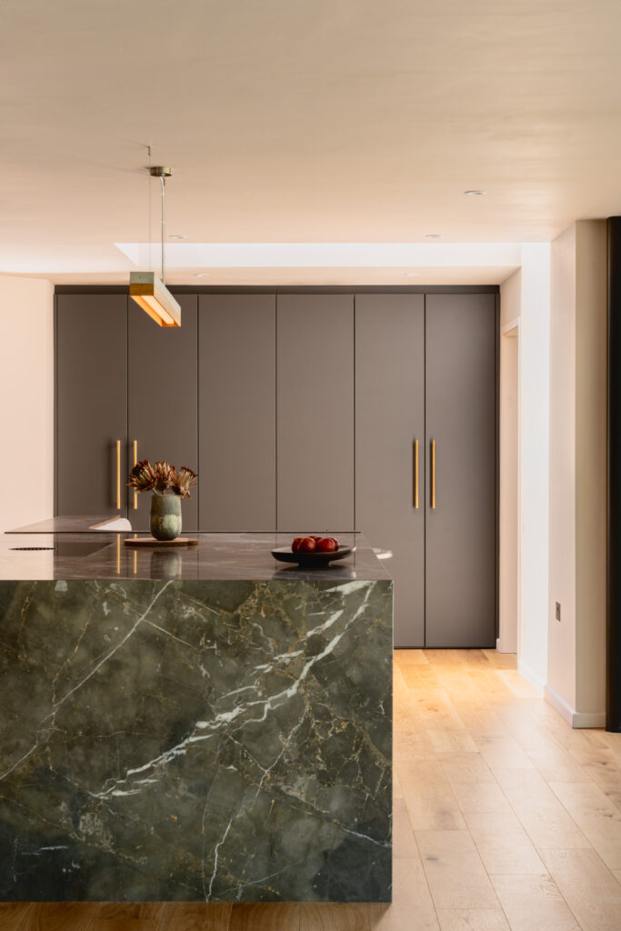 Modern kitchen with dark gray cabinets, brass handles, and a green marble island topped with a vase and fruit bowl.